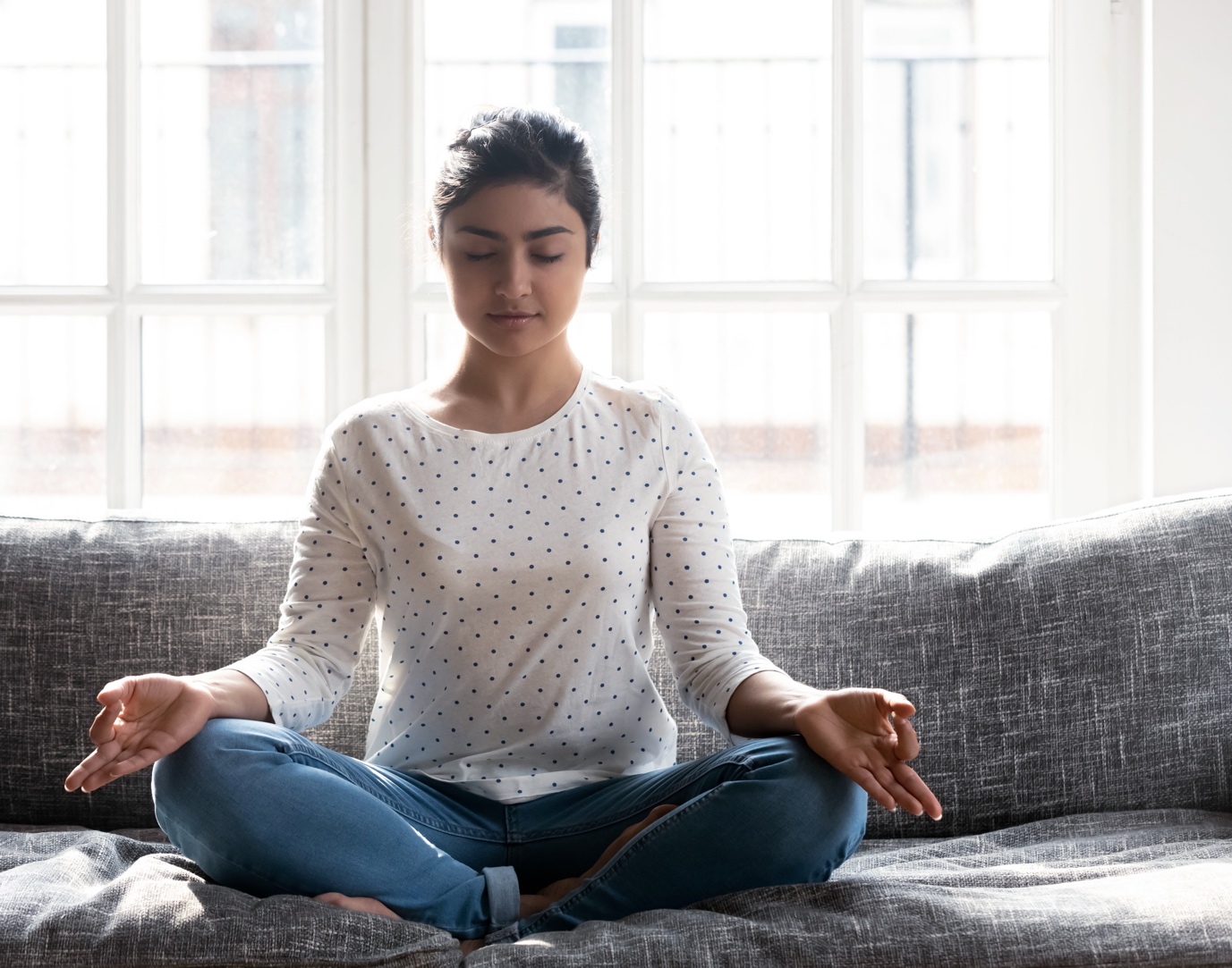 A woman meditating in a lotus pose on a gray sofa, with hands in gyan mudra, light streaming through large windows behind her.