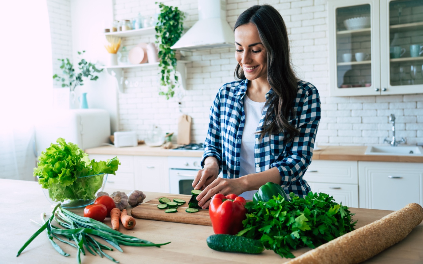 A woman chopping vegetables in a bright kitchen, smiling as she prepares a meal with fresh produce laid out on the counter.