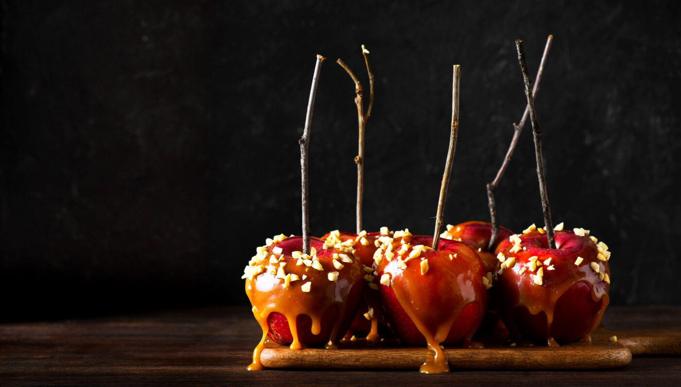 Caramel apples with nuts on wooden sticks, coated in dripping caramel sauce and sprinkled with chopped nuts, displayed on a wooden board against a dark background.